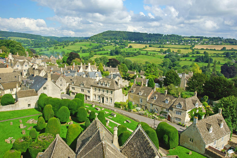 A photograph taken from a drone of a Cotswold village with rolling Cotswold hills in the background